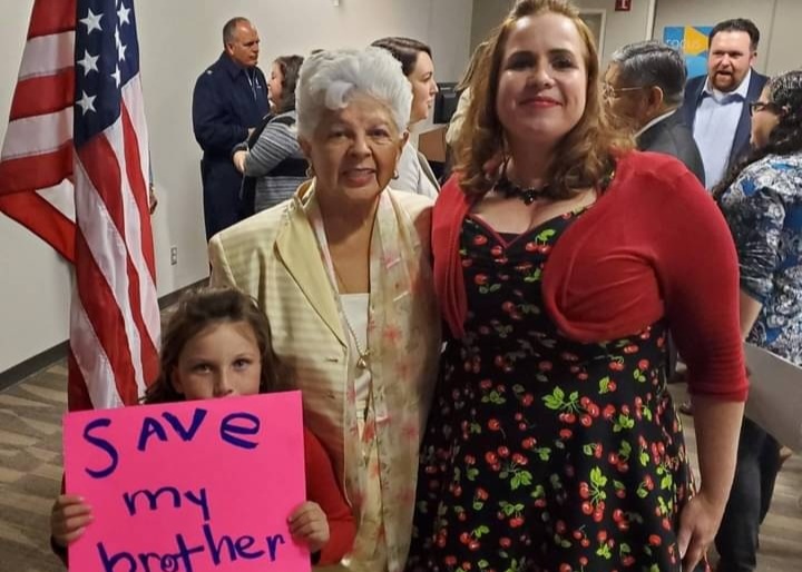 A young girl holds a pink sign that says "Save my brother" as two other women stand near. 