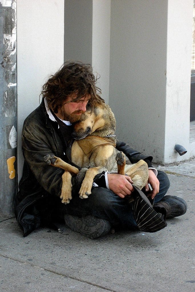 homeless man with longish brown hair and beard in old clothing sits on street closely cuddlinghis large tan-and-gray dog
