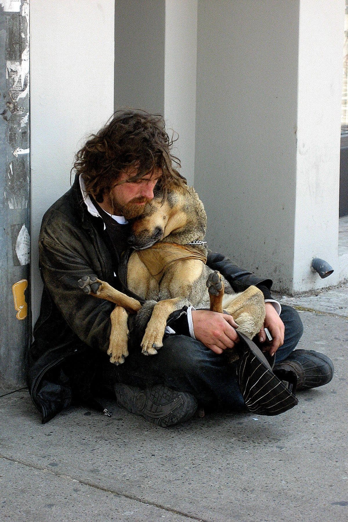 homeless man with longish brown hair and beard in old clothing sits on street closely cuddlinghis large tan-and-gray dog