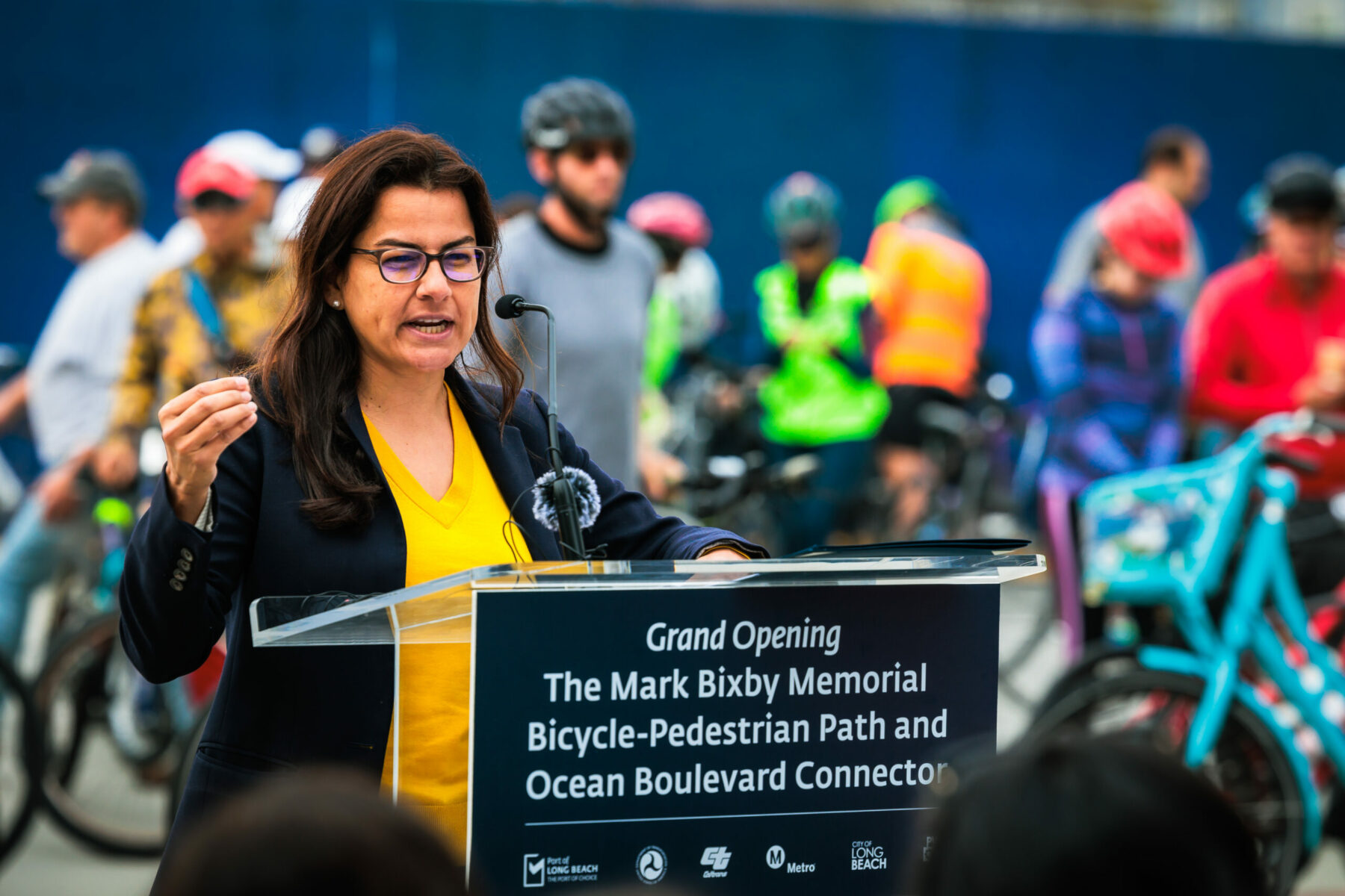 A woman speaks at a lectern that has sign reading, "Grand Opening The Mark Bixby Memorial Bicycle-Pedestrian Path and Ocean Boulevard Connector."