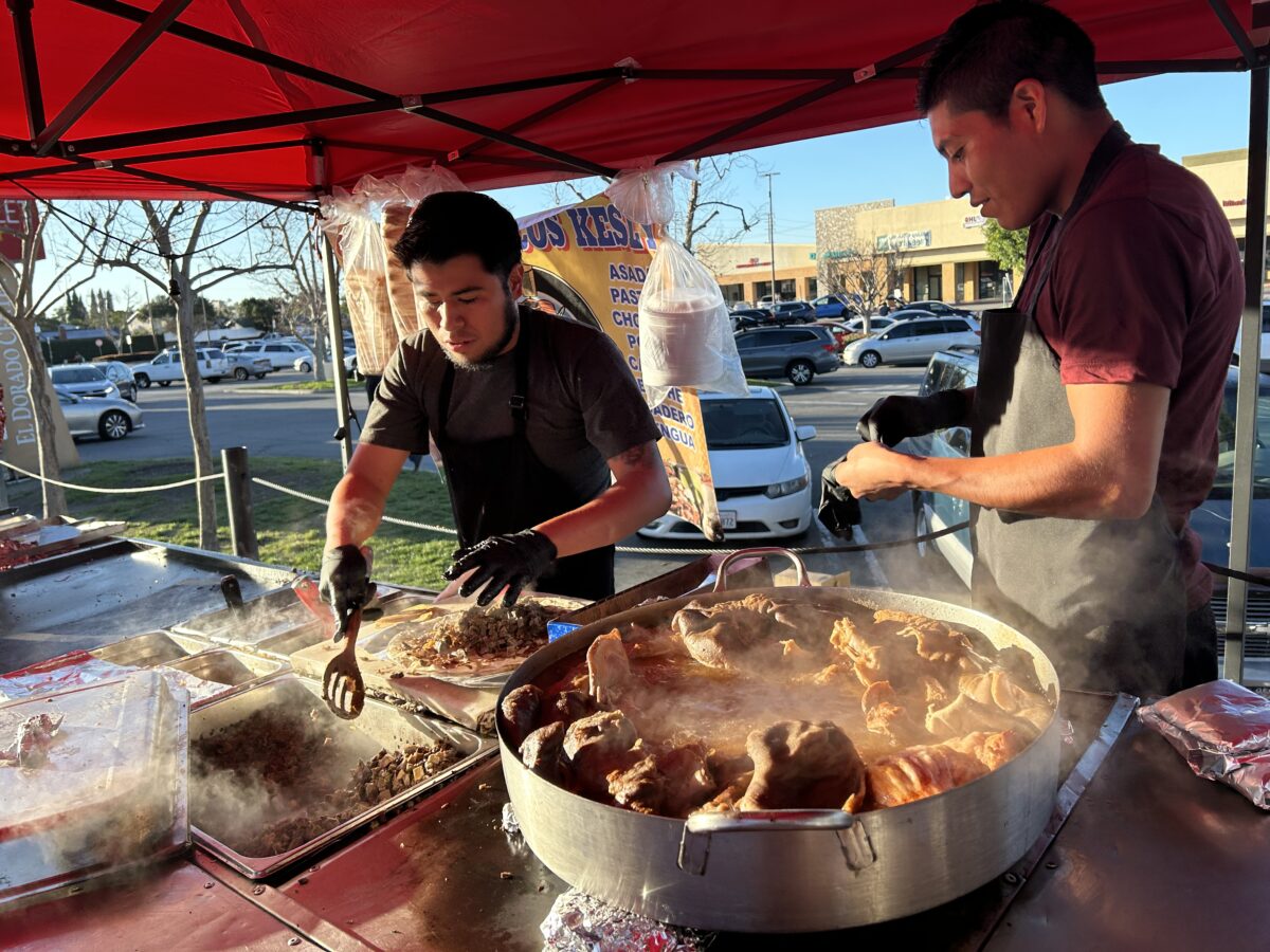 Two people cook tacos at a sidewalk stand.