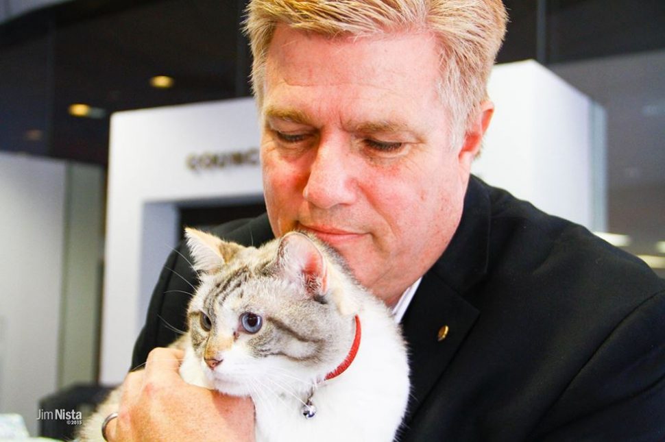 Blond man in black jacket holds big white cat with a gray striped head.