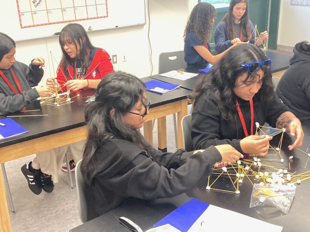 Three pairs of girls sitting at tables work on an engineering activity using spaghetti and marshmallows.